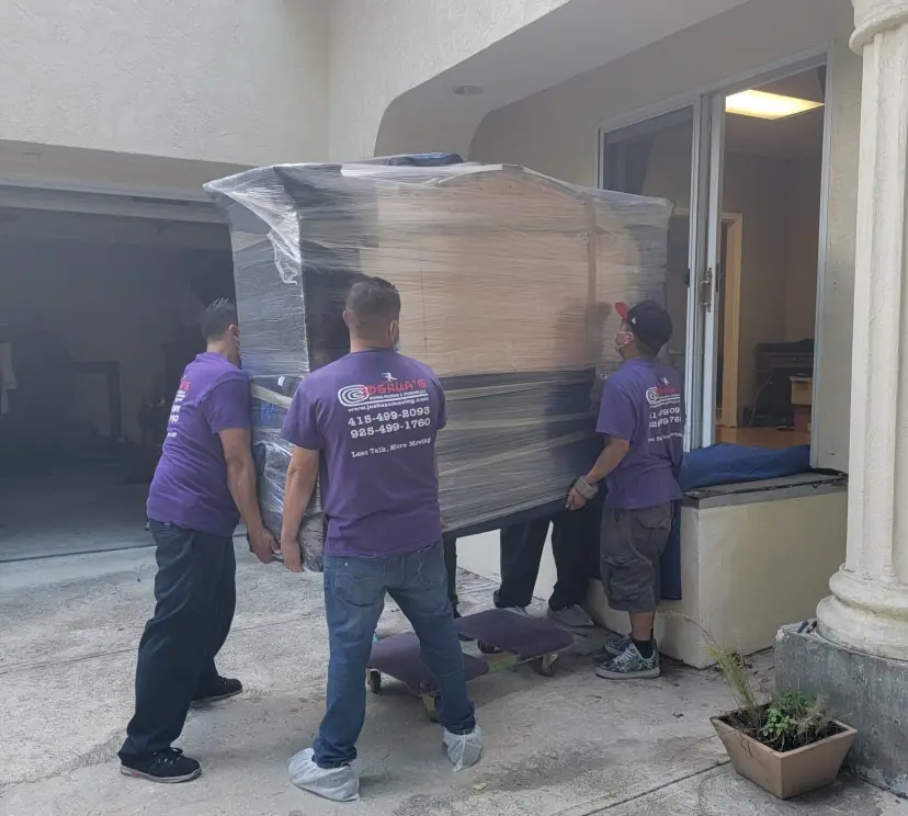Three movers carrying a plastic-wrapped dresser through the front entry of a house